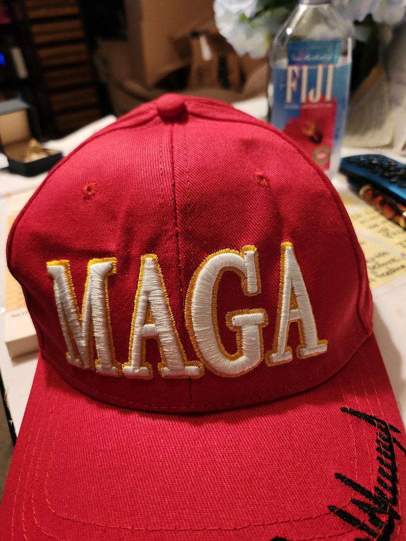 May include: Red baseball cap with a white and gold embroidered "MAGA" logo. The brim of the cap has a black embroidered design.