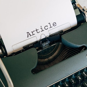 May include: An Olympia typewriter in a teal-green hue, with a sheet of paper reading "Article" inserted. The keyboard is black with white keys. The typewriter is on a white surface.