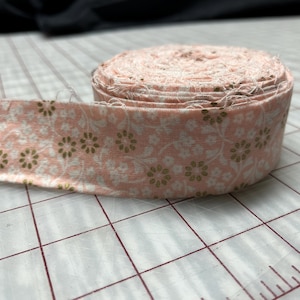 May include: A roll of fabric ribbon featuring a light peach background with a repeating pattern of small white floral outlines and metallic gold flower centers. The ribbon has frayed edges and is coiled into a circular shape on a flat surface.