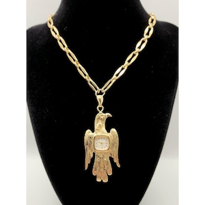 May include: A gold-toned necklace featuring a textured eagle-shaped pendant with an integrated analog watch face. The watch has a white dial with black numerals and thin gold hands. The pendant hangs from a thick, oval-linked gold-toned chain.
