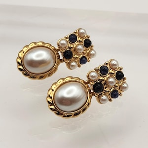 May include: A pair of gold-toned earrings. Each earring features a large, oval, faux pearl surrounded by a gold rope-like border. A cluster of small white, navy, and blue-toned stones adorns the top of each earring.
