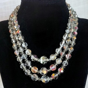 May include: A multi-strand necklace featuring clear, faceted beads. The beads have an iridescent quality, reflecting colors like pink, orange, and yellow. The necklace is displayed on a black background, highlighting the sparkle of the beads.