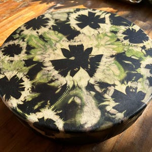 May include: A circular drum featuring a black and light green marbled pattern across its surface. The design consists of dark, solid black floral-like shapes interspersed with swirling, light green and off-white paint textures.
