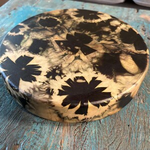 May include: A circular wooden disc featuring a high-gloss resin finish with a black and cream-colored floral pattern. The design displays dark, solid black flower shapes contrasted against a swirling, marbled cream and light brown wood grain background.