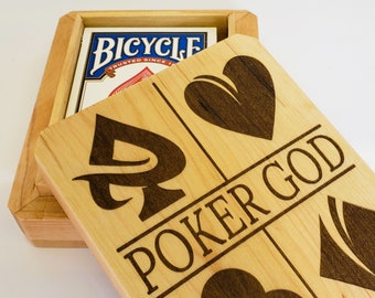 May include: A wooden box with a hinged lid. The lid is open and shows a design of four playing card suits, a spade, a heart, a club, and a diamond. The text "Poker God" is engraved on the lid. The box is made of light wood and has a natural finish.