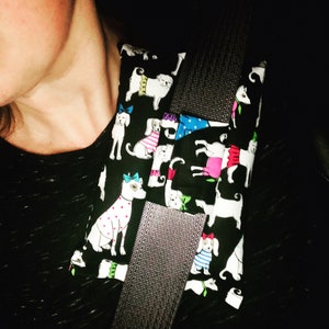 May include: A black fabric car seatbelt cover with a pattern of white dogs wearing colorful outfits. The dogs are wearing hats, dresses, and bow ties.