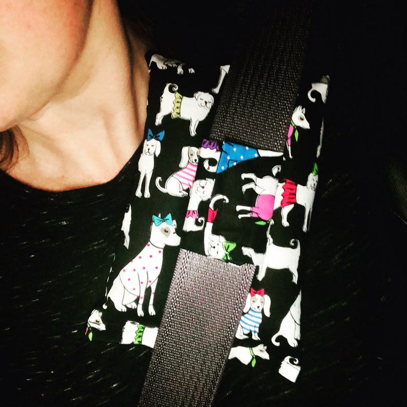 May include: A black fabric car seatbelt cover with a pattern of white dogs wearing colorful outfits. The dogs are wearing hats, dresses, and bow ties.