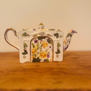 May include: A white porcelain teapot with a floral design. The teapot features a rectangular shape with a curved handle and spout. The front of the teapot has a painted scene of yellow flowers and purple accents. Gold trim accents the edges and lid.