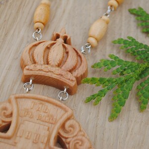 May include: A wooden pendant necklace featuring a carved crown and cross. The cross has the words "I AM THE WAY" inscribed on it. The necklace is made from wooden beads and silver chains.
