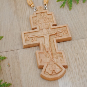 May include: A wooden cross with intricate carvings and a figure of Jesus Christ on the front. The cross is made of light brown wood and has a beaded chain attached to the top.