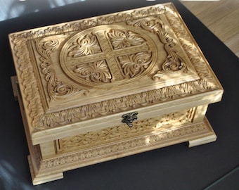 May include: A handcrafted wooden box with intricate carvings. The box features a detailed cross design on the lid, surrounded by floral patterns. The natural wood color and ornate details give it a vintage aesthetic. The box is rectangular with a hinged lid and metal clasp.