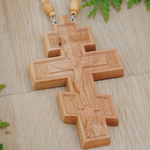 May include: A wooden cross with intricate carvings, including a depiction of Jesus Christ on the cross. The cross is attached to a brown cord with wooden beads.
