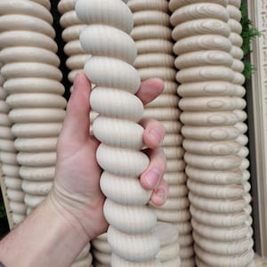 Op de afbeelding: Een lichtgekleurde, onafgewerkte houten spil met een spiraalvormig ontwerp. De spil wordt in een hand gehouden, met verschillende soortgelijke spillen op de achtergrond gestapeld. De houtnerf is zichtbaar en de algehele esthetiek is rustiek.