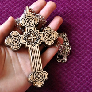 May include: A wooden cross pendant featuring intricate carved detailing and a clover-like shape at each end, suspended from a silver-toned metal chain. The wood displays a natural grain pattern with dark stained accents in the recessed areas.