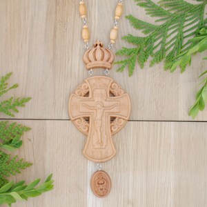 May include: A wooden cross with a carved image of Jesus Christ on the front. The cross is on a beaded chain with a crown at the top and a small round pendant at the bottom.