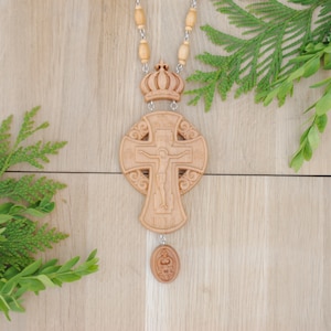 May include: A wooden cross with a carved image of Jesus Christ on the cross. The cross is decorated with intricate carvings and is attached to a wooden chain with a crown at the top. The cross is hanging against a light brown wooden background.
