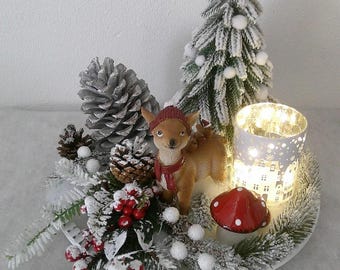 Könnte beinhalten: Ein dekoratives Tischgesteck mit einer braunen Rehfigur mit roter Mütze und Schal, einem beschneiten Miniatur-Tannenbaum, einem silberfarbenen Tannenzapfen, roten Beeren, einem kleinen roten Fliegenpilz und einem zylindrischen Windlicht.