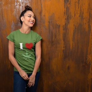 May include: Olive green t-shirt with the text "I ♥" and a cannabis leaf graphic. The heart is red, and the "I" is white. The shirt is worn with blue jeans. The background is a textured, rusty brown wall.