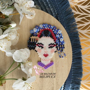 Può includere: Un ritratto in perline di una donna con capelli neri, labbra rosse e un copricapo blu e oro. Il ritratto è realizzato con piccole perline ed è montato su uno sfondo di legno. Il testo "LE BAZAR DE MILIPUCE" è visibile sotto il ritratto.