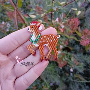 May include: A beaded deer figurine with a festive design. The deer is brown with white spots, a red and white hat, and a green and red wreath. The text "LE BAZAR DE MILIPUCE" is visible.