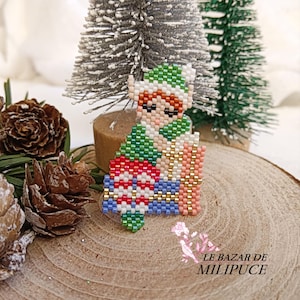 May include: A beaded Christmas elf ornament. The elf is wearing a green and white hat and outfit, holding a red and white gift. The ornament is on a wooden surface with small Christmas trees in the background.