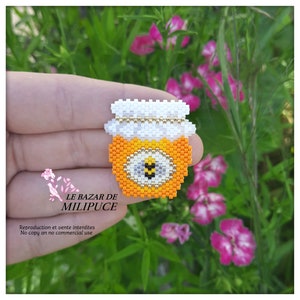 Peut inclure: Un petit pot de miel en perles avec un couvercle blanc et un motif d'abeille jaune et noir. Le pot est tenu dans une main sur un fond floral vert et rose.