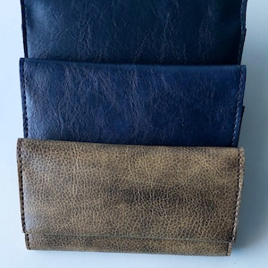 Puede incluir: Tres carteras de cuero en tonos azules y marrones. La cartera superior es azul marino oscuro, la del medio es azul más claro y la inferior es marrón texturizado. Todas las carteras son rectangulares con un diseño plegado.