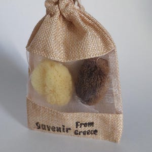 May include: A small burlap bag with a drawstring closure contains two natural sea sponges. The bag is translucent and has the text "Souvenir From Greece" printed on the front.