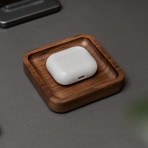 May include: A white wireless earbud case rests in a small, square, wooden tray. The tray is made of dark brown wood with a raised edge. The background is a neutral gray surface, with a phone stand and other objects visible.