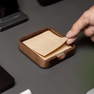 May include: A wooden memo pad holder with a stack of graph paper. The holder is rectangular with a dark brown finish. A hand is pulling a sheet of paper from the pad. Other items on the desk include a phone, laptop, and mouse.