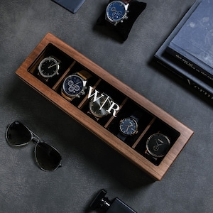 May include: A wooden watch box with six slots for storing watches. The box is made of dark wood and has a clear glass top. The box is labelled "WTR" on the front. There are five watches in the box, all with different styles and colours.