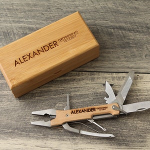 May include: A wooden box with the text "ALEXANDER GROOMSMAN 10.13.2017" engraved on the top. The box contains a multi-tool with a wooden handle and the same text engraved on it.