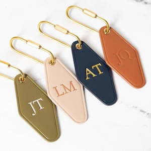 May include: Four diamond-shaped keychains with gold-toned metal loops. The keychains are olive green with JT, light pink with LM, navy blue with AT, and terracotta with JQ embossed in gold or white lettering. Each tag has a smooth, matte finish.