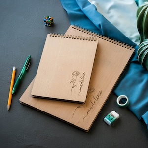 May include: Three wooden spiral-bound notebooks with the name "madeline" and a floral design engraved on the cover. A pencil, green pen, Rubik's cube, decorative green cacti, and washi tape are also present.