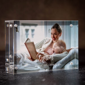 May include: A clear acrylic photo block displays a photo of a mother reading to a child. The photo block has the word "Mom" and the phrase "All that I am, I owe to you" etched on the front. The photo block is rectangular and has silver screws.
