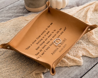May include: A tan leather tray with a diamond-shaped design and rolled edges. The tray has a quote and names engraved in black. Two silver rings are inside the tray. The text reads "Like a river flows surely to the sea...ANNA & KURT 03.05.2019."