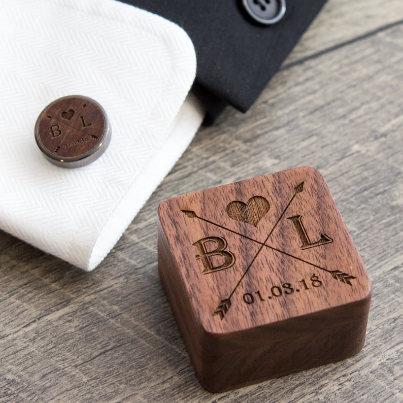 Puede incluir: Una caja de madera con un coraz&oacute;n, flechas y las iniciales "B L" grabadas en la parte superior. La fecha "01.08.18" tambi&eacute;n est&aacute; grabada en la caja. Un gemelo a juego con el mismo dise&ntilde;o se muestra en un pu&ntilde;o de camisa blanca.