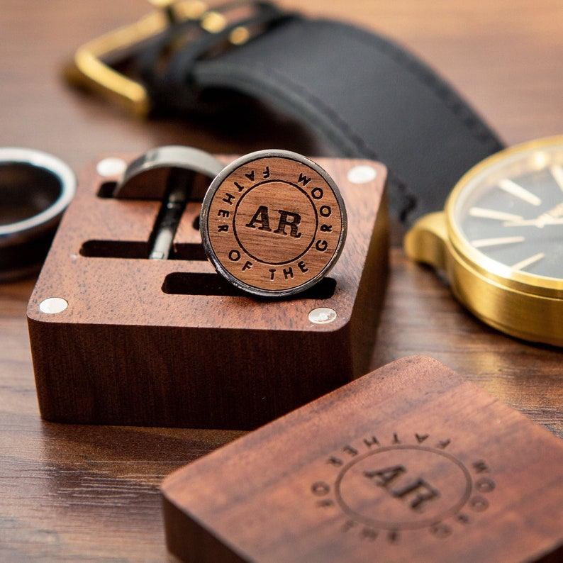 Puede incluir: Una caja de madera con un acabado marr&oacute;n oscuro contiene dos gemelos. Los gemelos son redondos y est&aacute;n hechos de metal con una incrustaci&oacute;n de madera. La incrustaci&oacute;n presenta el texto "Father of the Groom" y las iniciales "AR".