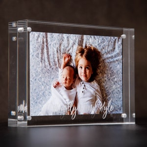 May include: A clear acrylic photo frame with a picture of two children, a baby and a toddler, lying on a patterned blanket. The text "Charlie and Kyle" is written on the bottom of the photo.