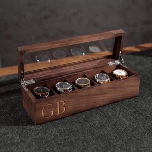 May include: A wooden watch box with a clear glass lid, displaying five watches. The box is engraved with the initials "GB". The watches have various band and face colors. The box is on a dark gray surface.