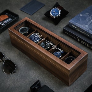 May include: A wooden watch box with a glass top and six compartments for storing watches. The box is engraved with the name "Ashley Gregory".