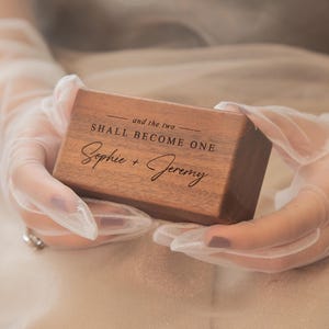 May include: A rectangular wooden ring box with a dark brown finish. The top surface is engraved with the text "and the two SHALL BECOME ONE" followed by the names "Sophie + Jeremy" in a combination of serif and script fonts.