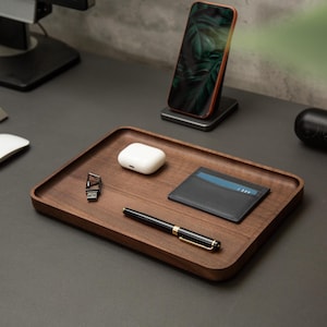 May include: A rectangular dark wood valet tray holding a black leather card holder, a black and gold fountain pen, a white wireless earbud charging case, and a small silver USB flash drive with a geometric metal casing.