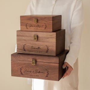 May include: Three wooden boxes of varying sizes are stacked, held by a person. Each box is a rich brown color, with a brass clasp and engraved text. The top box reads "Tyler & Abigail", the middle box reads "Samantha", and the bottom box reads "James & Sabrina".