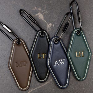 May include: Four diamond-shaped leather keychains with black metal carabiners. The keychains are brown with MD, black with gold LT, navy blue with silver AW, and green with gold LH embossed initials. Each features white contrast stitching.