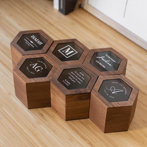 May include: Six hexagonal wooden boxes with clear glass tops. Each box has a different inscription, including names, initials, and dates. The boxes are arranged in a honeycomb pattern.