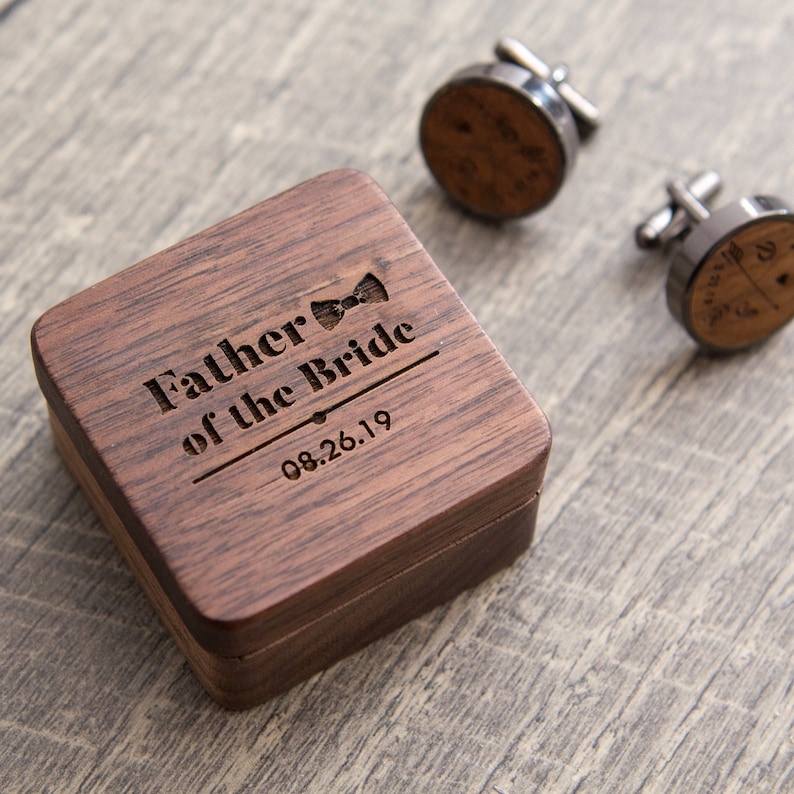 Puede incluir: Una caja de madera con la inscripci&oacute;n "Father of the Bride" y la fecha "08.26.19" grabada en la parte superior. Tambi&eacute;n se muestran dos gemelos de plata con un dise&ntilde;o de pajarita.