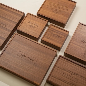 May include: A collection of wooden boxes with engraved names and dates. The boxes are various sizes and shapes, with dark brown wood grain. Names like "Steven & Venus" and "Thomas & Melissa Hernandez" are visible.