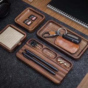 May include: A collection of wooden desk organizers, including trays and a notepad holder. The trays hold items like pens, keys, paperclips, and SD cards. The items are arranged on a dark gray felt mat and a wooden desk.