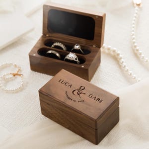 May include: Two rectangular dark brown wooden ring boxes. One box is open, revealing a black interior with four slots holding silver-toned rings. The other box is closed, featuring the engraved text LUCA & GABE and NOVEMBER 10, 2026.